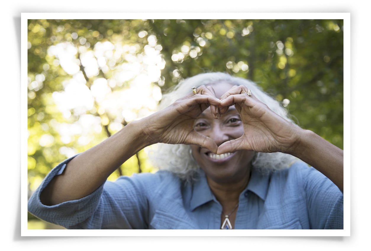 A senior woman with gray and white hair in the park.  She's smiling and making the heart shape with her hands.  She's  looking at the camera through the heart.  She is wearing a blue shirt and undershirt.  She is African ethnicity.
